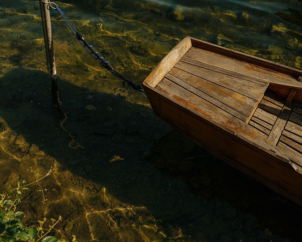 Holzboot (Plätte) angebunden an Metallpfahl im Wasser, ruhige Atmosphäre.