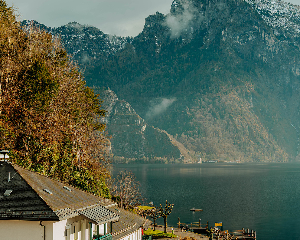 Traunsee mit der „Schlafenden Griechin“ im Hintergrund, weißes Haus mit Flaggen, Holzstege am Ufer.